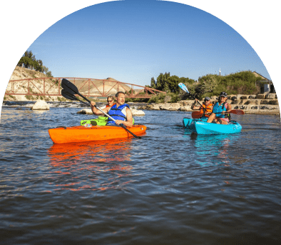Two people kayaking on a calm river near rocky cliffs under a clear sky.