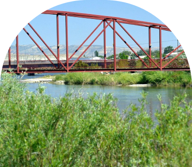 Red steel bridge over a calm river with greenery in the foreground.